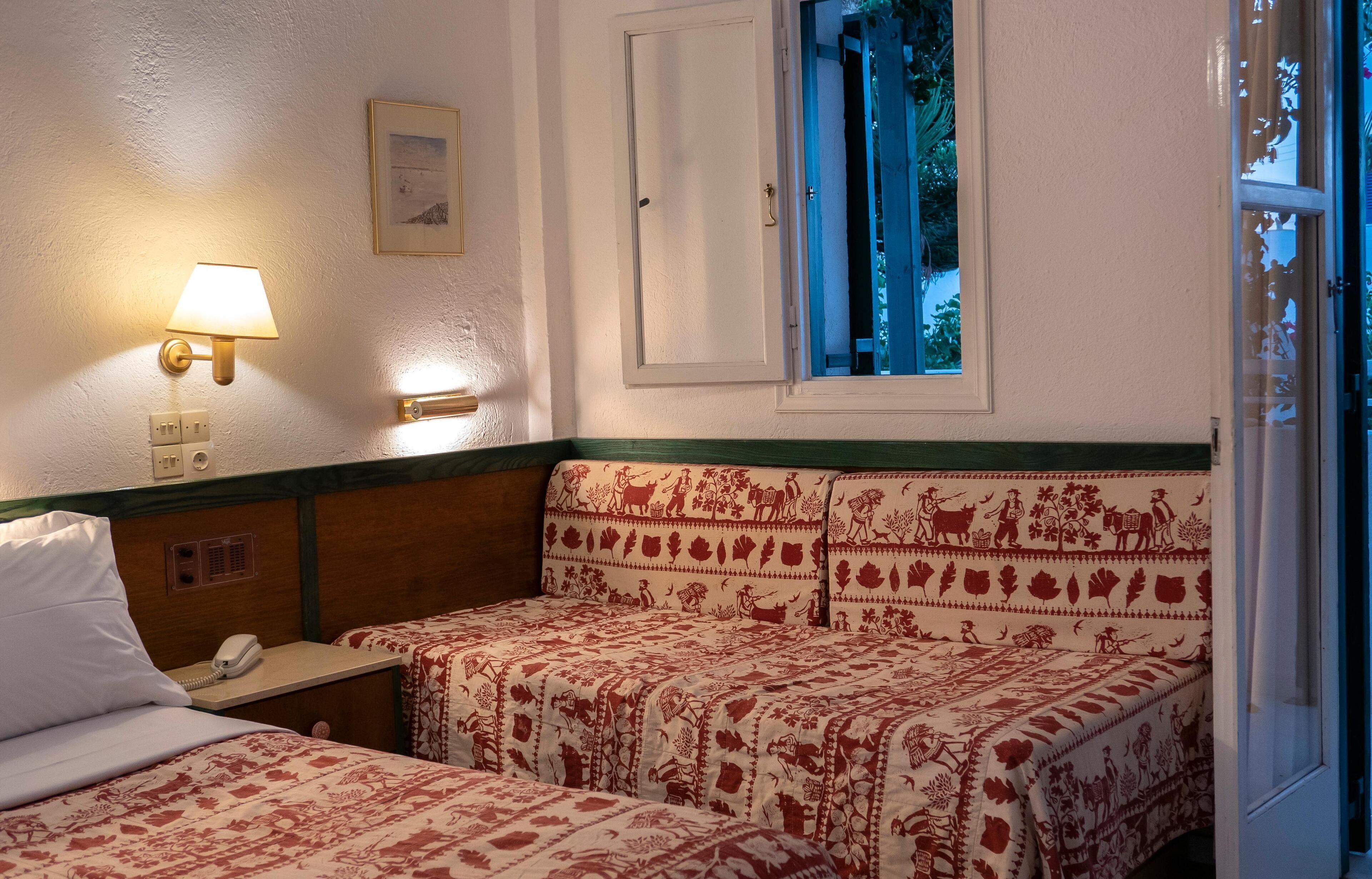 A room with two beds, a bedside table with a lamp, a phone, and a window with a view of greenery.
