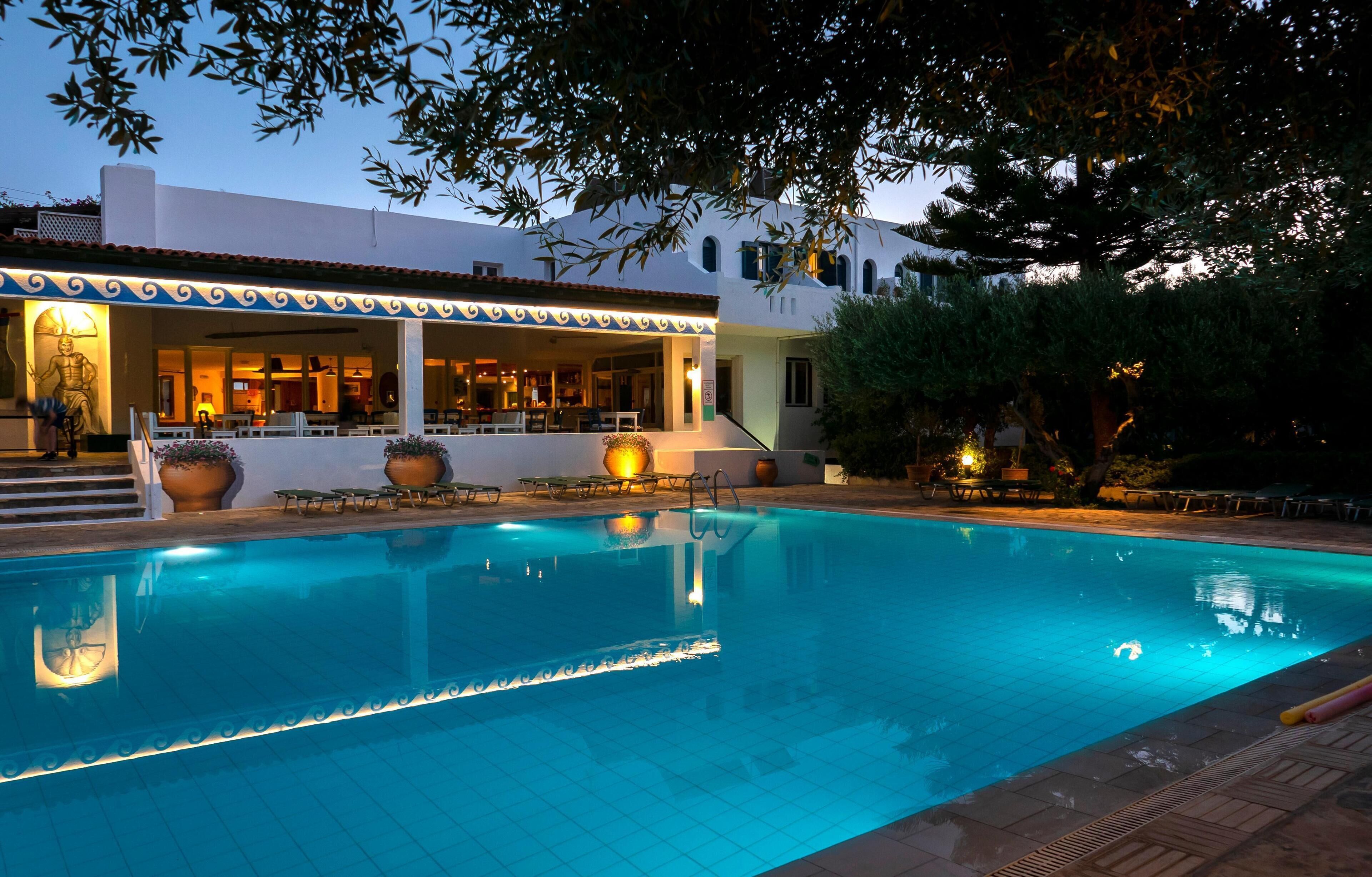 A swimming pool with a tiled deck, a covered seating area, and a building with a white facade and blue trim.