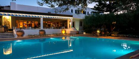 A swimming pool with a tiled deck, a covered seating area, and a building with a white facade and blue trim.