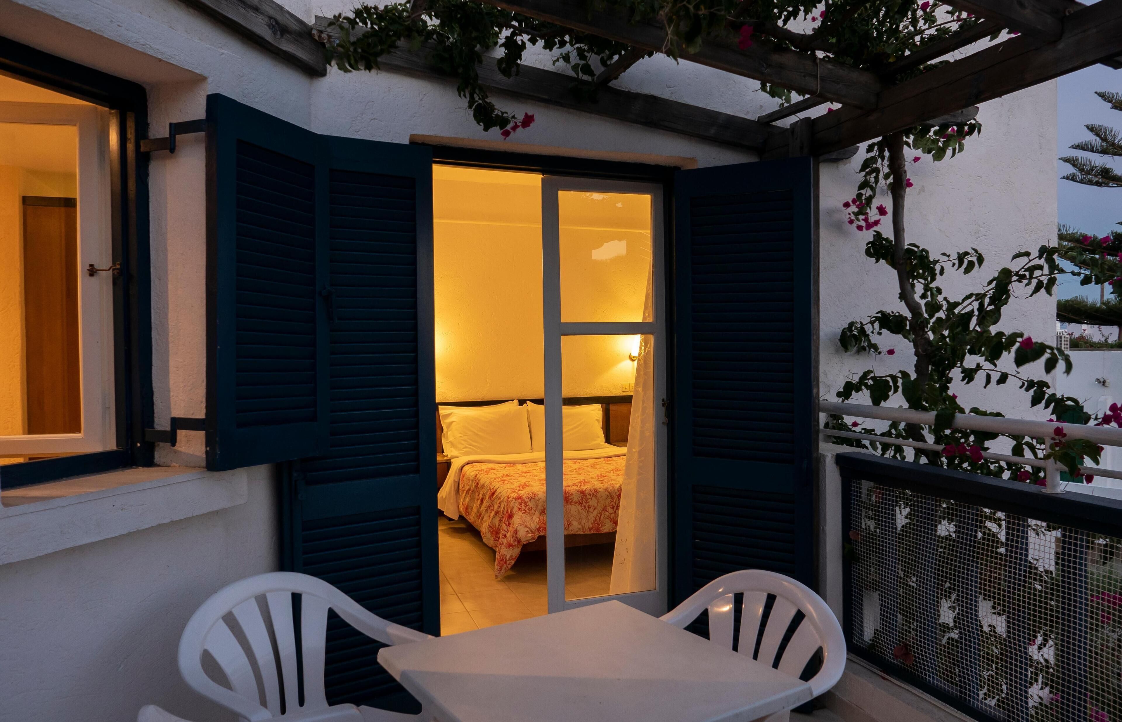 Ein Zimmer mit einem Bett, einem Fenster mit Rollläden und einem Balkon mit Stuhl und Tisch.