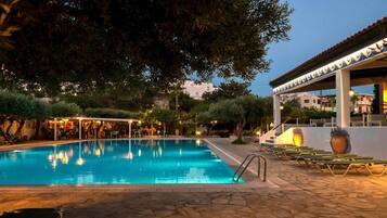 A swimming pool with a stone deck, surrounded by trees and a covered seating area with tables and chairs.