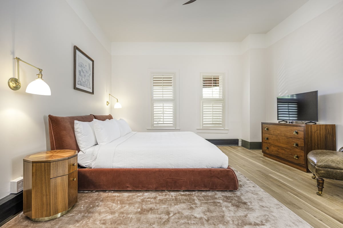 Une chambre à coucher comprenant un lit, des tables de chevet, une commode, un téléviseur et deux fenêtres équipées de stores.