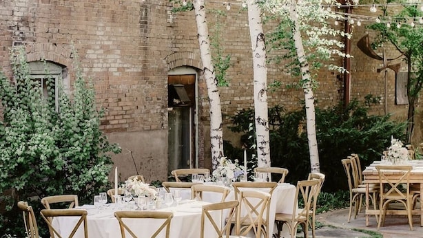 Un espace de restauration en plein air avec des tables disposées pour accueillir six personnes, entouré de bouleaux et bordé d’un bâtiment en briques.