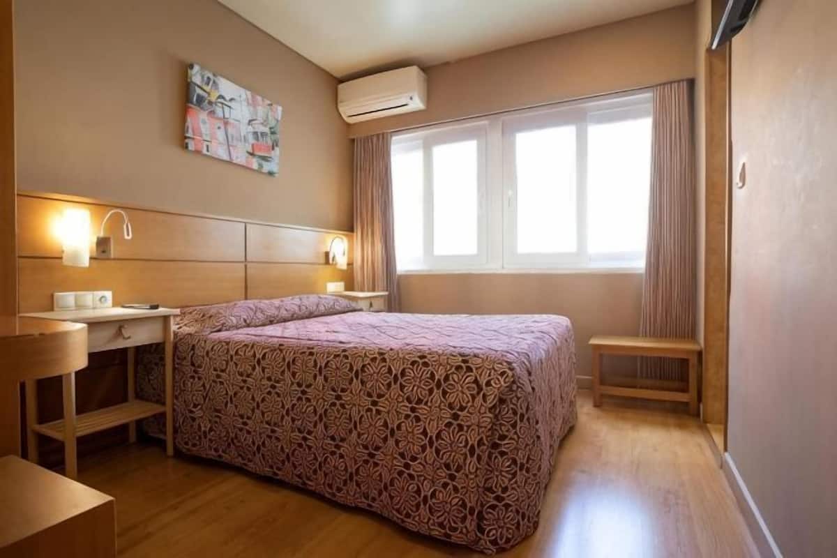 A hotel room with a bed, bedside table, lamp, and a window with curtains.
