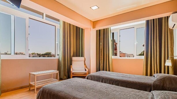A hotel room with two beds, a chair, a small table, and large windows with curtains.