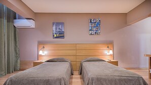 A hotel room with two single beds, a wooden headboard, a wall-mounted air conditioner, and two bedside tables with lamps.