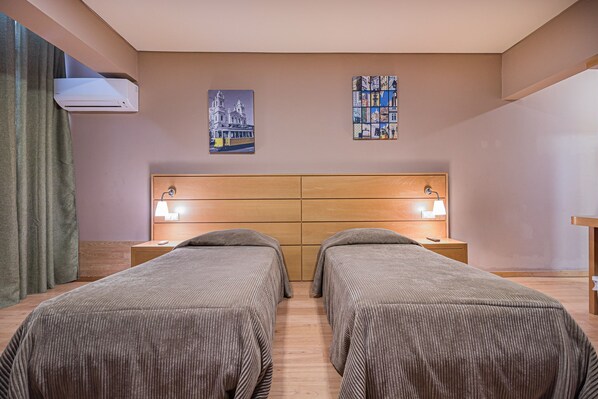 A hotel room with two single beds, a wooden headboard, a wall-mounted air conditioner, and two bedside tables with lamps.