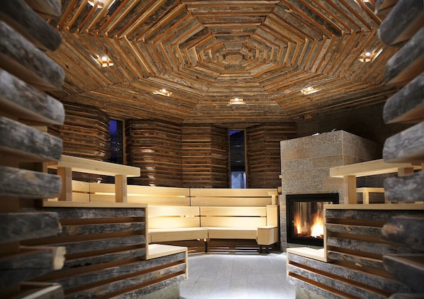A log cabin interior with a central fireplace, wooden benches, and a hexagonal ceiling design. - Tschuggen Grand Hotel (Arosa)