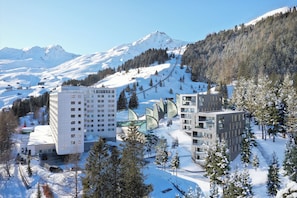 A modern hotel complex with multiple buildings, surrounded by snow-covered mountains and trees.