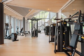 A gym with cardio machines, weight stations, and a large window providing natural light. - Tschuggen Grand Hotel (Arosa)