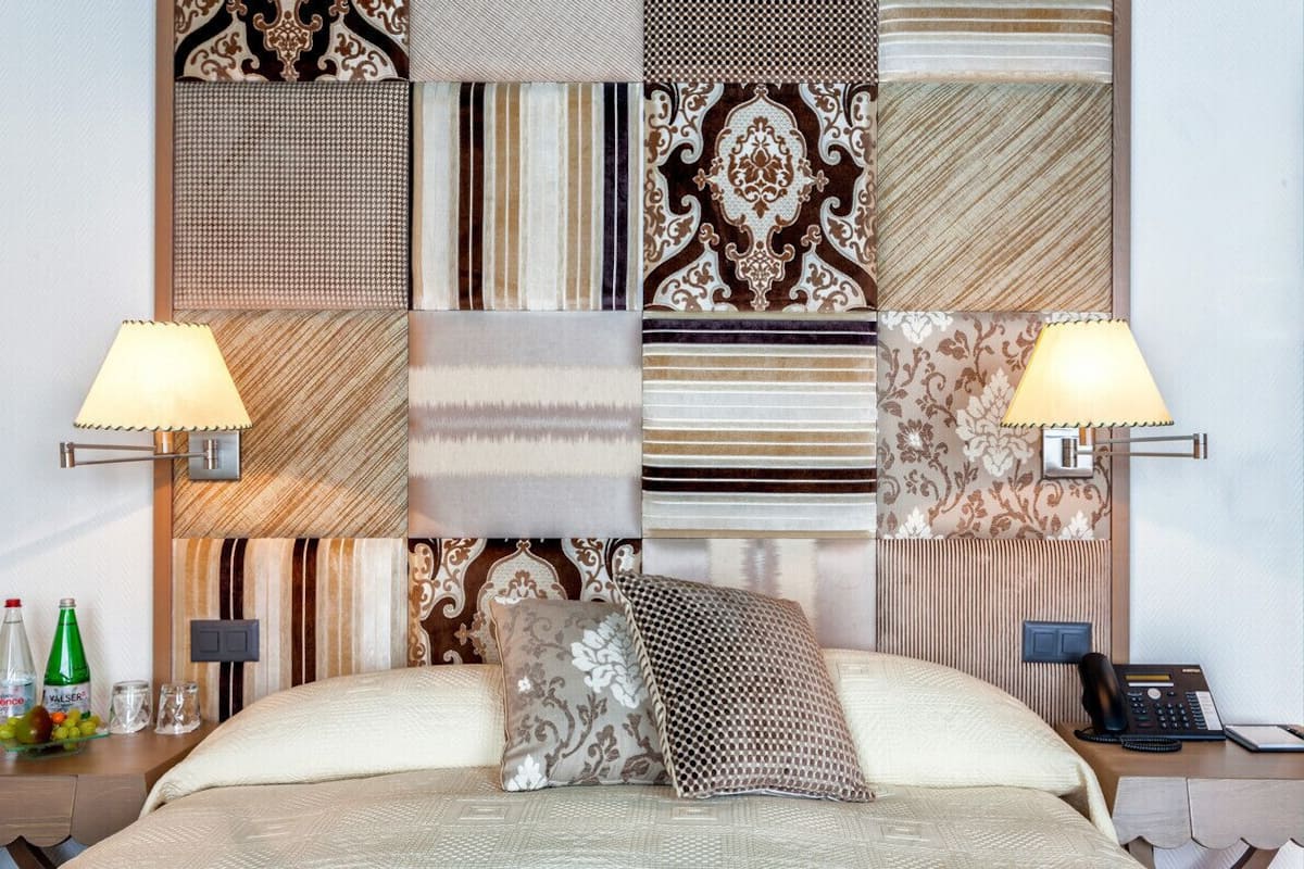 A hotel room with a large bed, two bedside lamps, a nightstand with a phone and a bottle of water, and a wall with various patterned wallpapers.
