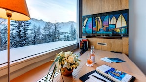 A hotel room with a large window, a wooden desk, a vase of flowers, a glass of orange juice, and a painting of colorful windows.