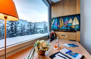 A hotel room with a large window, a wooden desk, a vase of flowers, a glass of orange juice, and a painting of colorful windows. - Tschuggen Grand Hotel (Arosa)