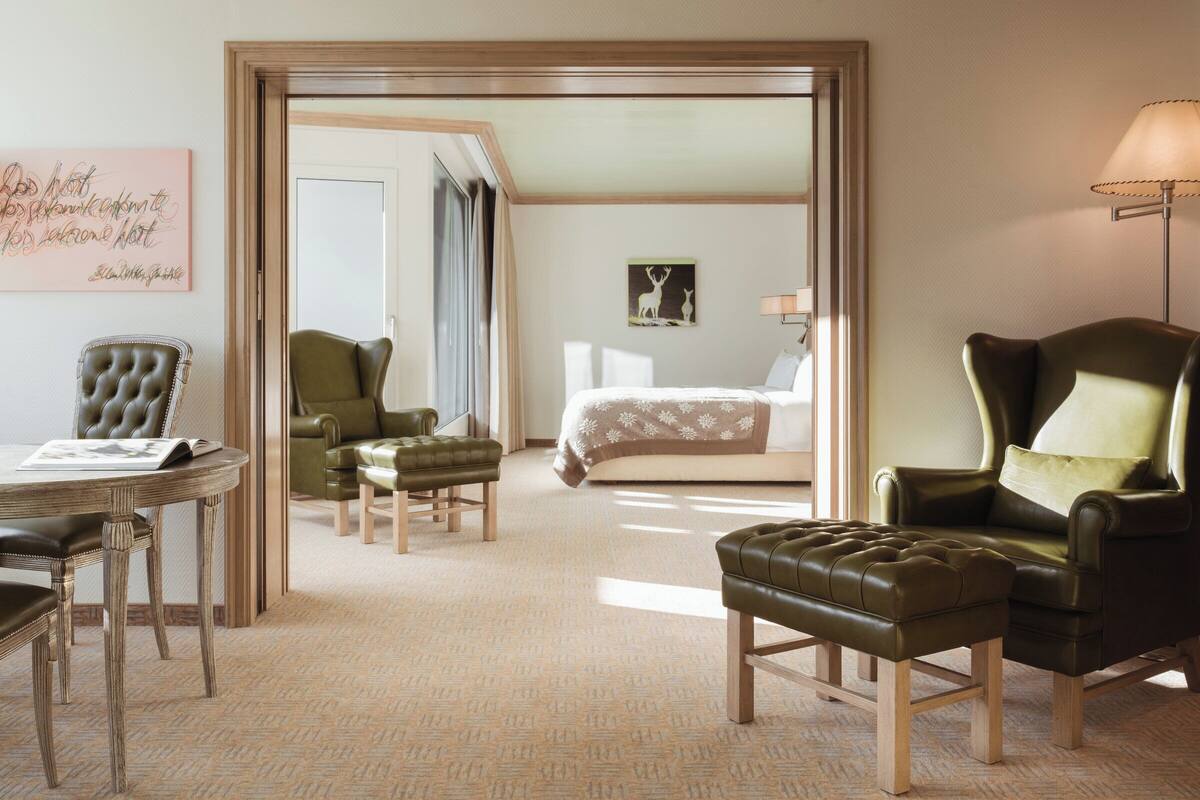 A hotel room with a bed, two green armchairs, a leather armchair, a wooden table with a magazine, and a patterned carpet.