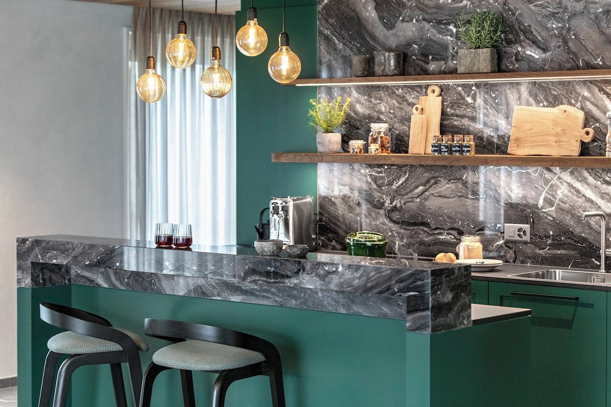 A modern kitchen with a marble island, bar stools, hanging light fixtures, and a green backsplash.