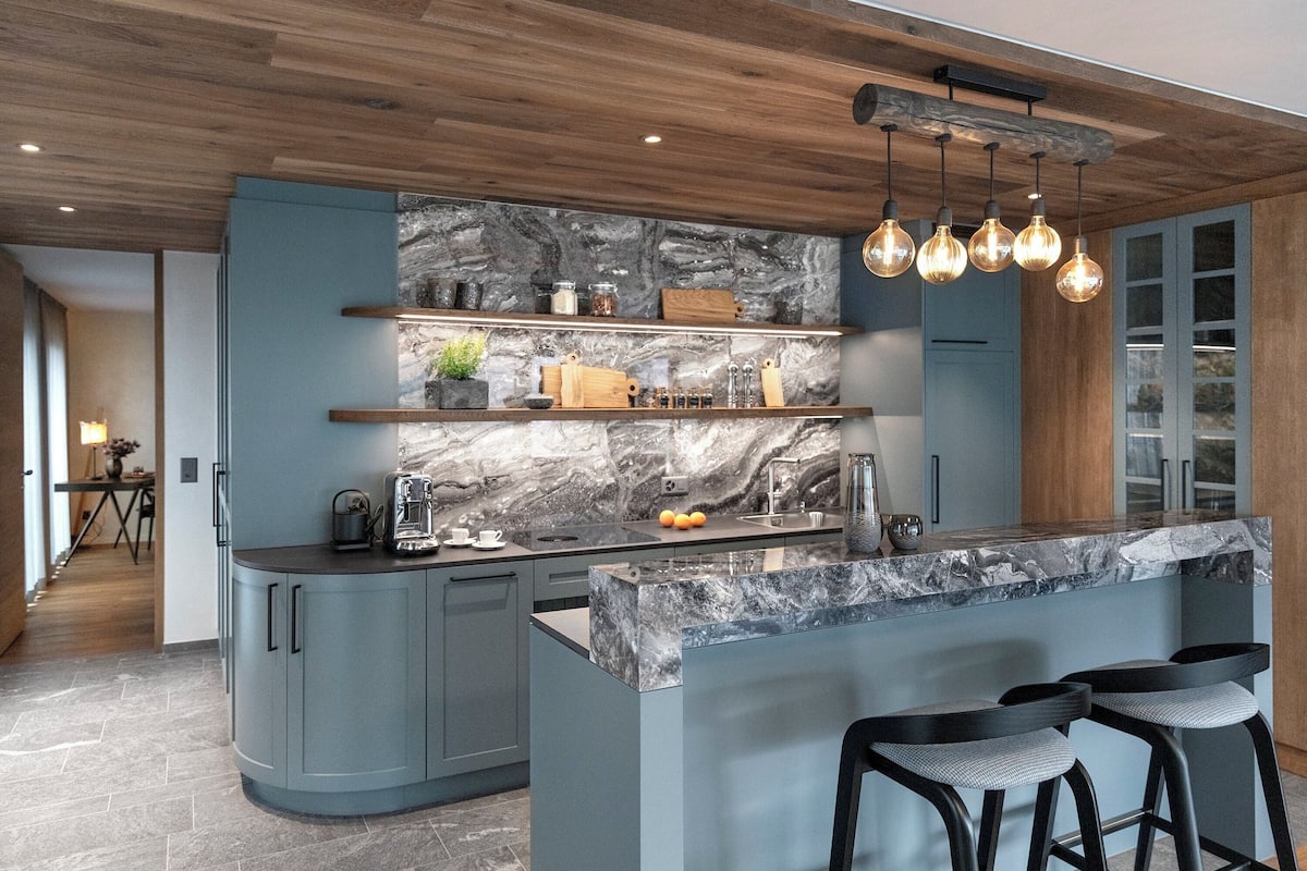 A modern kitchen with a marble island, bar stools, and a wooden ceiling.
