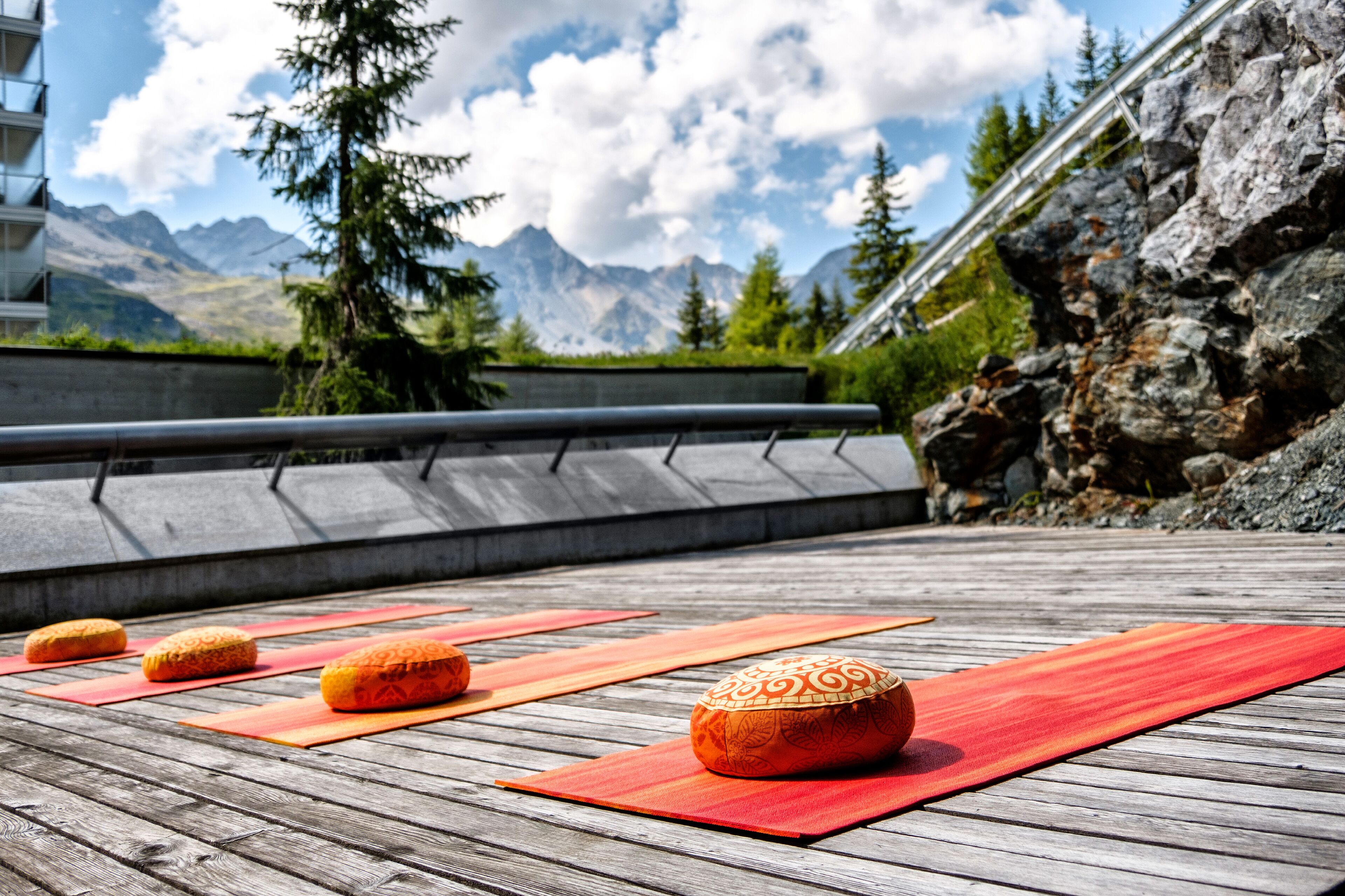 A wooden deck with yoga mats and decorative pillows, overlooking a mountainous landscape with a building in the background.