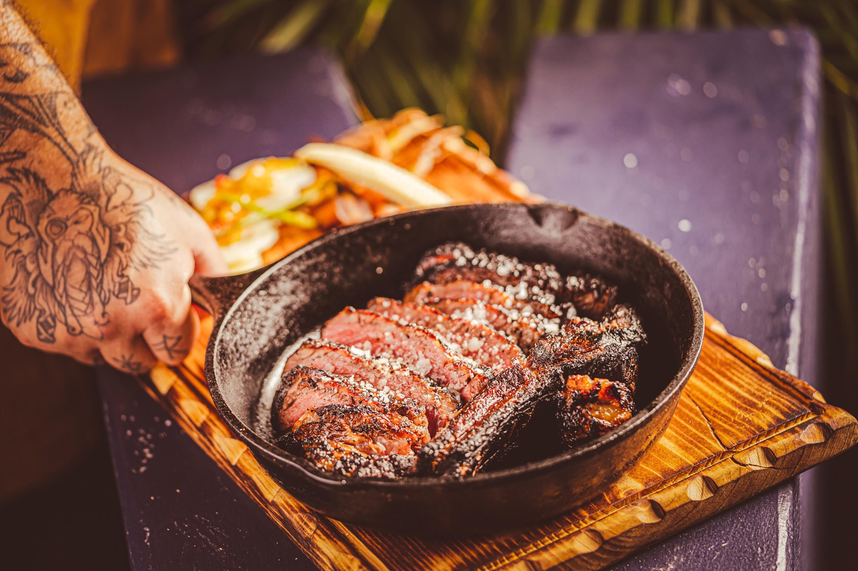 A steak in a skillet on a wooden board with a hand holding it.