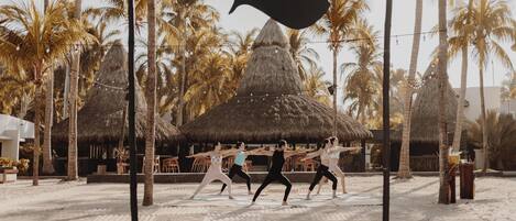 A beach with palm trees, a thatched-roof hut, and people exercising.