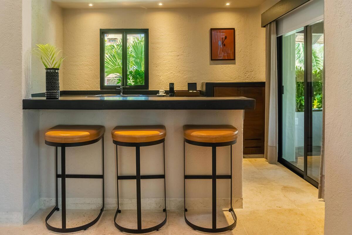 A kitchen with a black countertop, three stools, a window with greenery, and a framed picture on the wall.