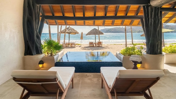 Zona de piscina con sillones de descanso, vista a la playa y sombrillas de paja.