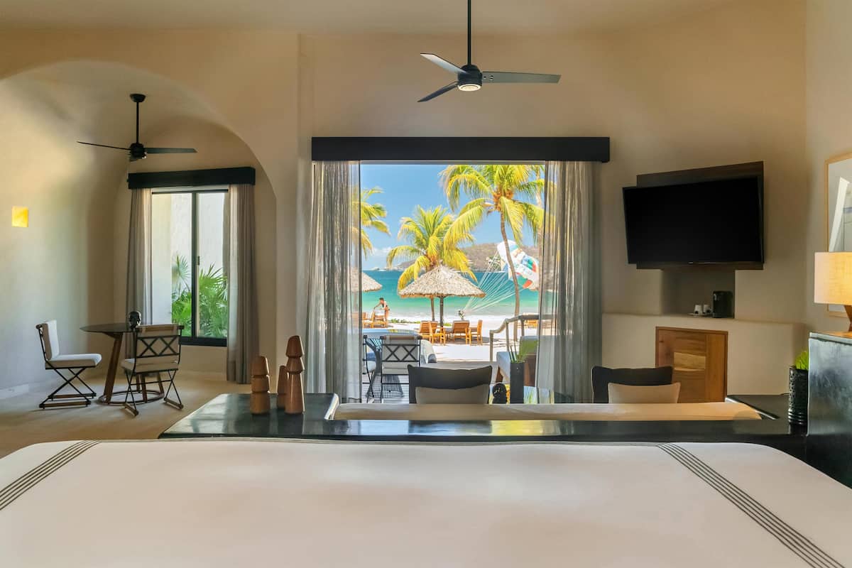 A hotel room with a bed, a TV, a ceiling fan, a window with a view of a beach, and a dining area with chairs and a table.