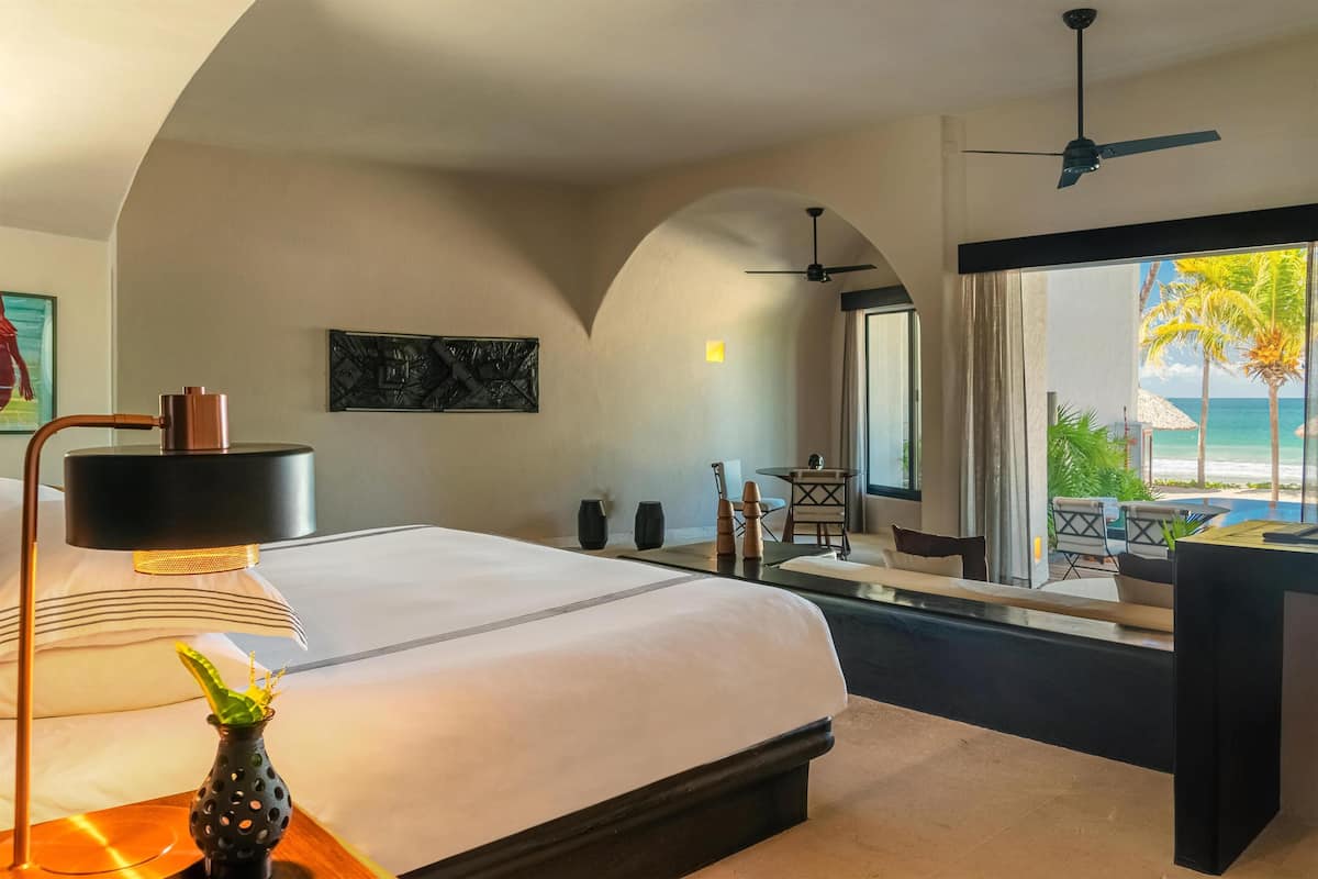 A hotel room with a bed, a desk, a chair, a vase with a plant, a ceiling fan, and a view of the ocean.