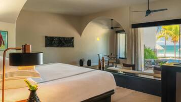 Habitación de hotel con cama, escritorio, silla, un florero con planta, ventilador de techo y vista al océano.