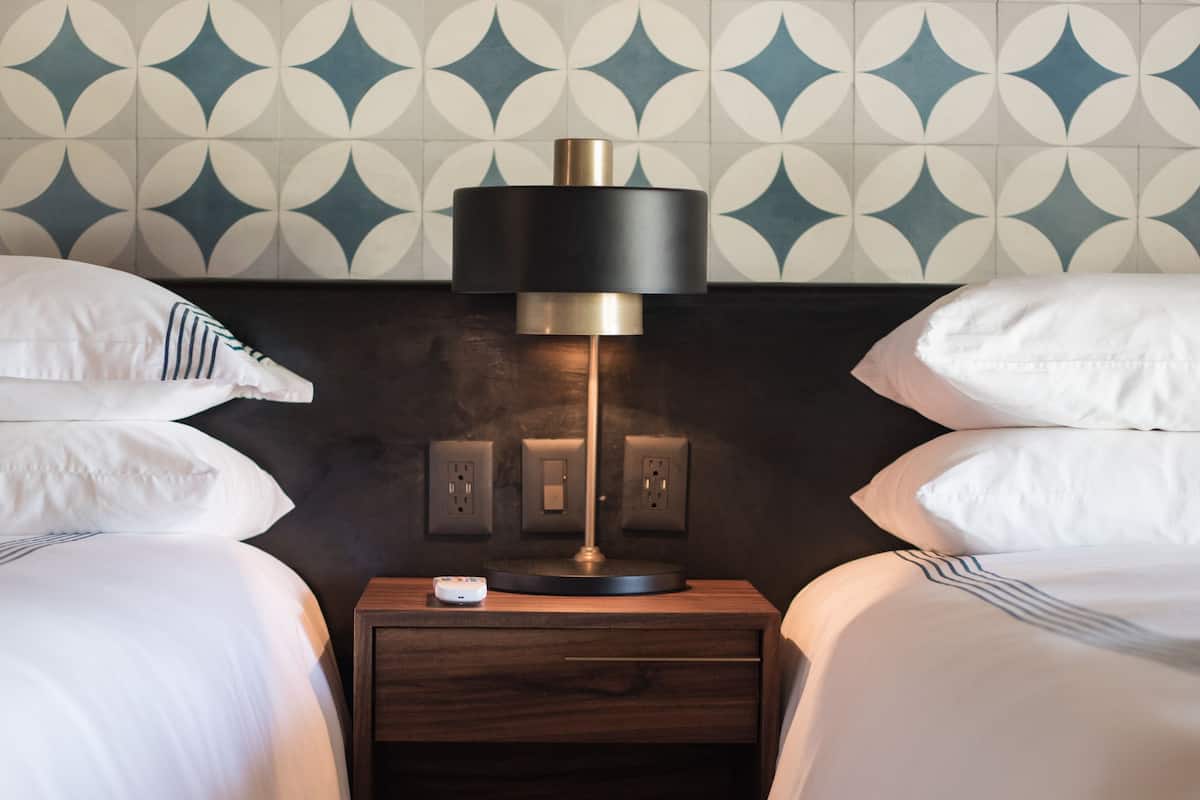 A hotel room with two beds, a nightstand with a lamp, and a patterned wall.