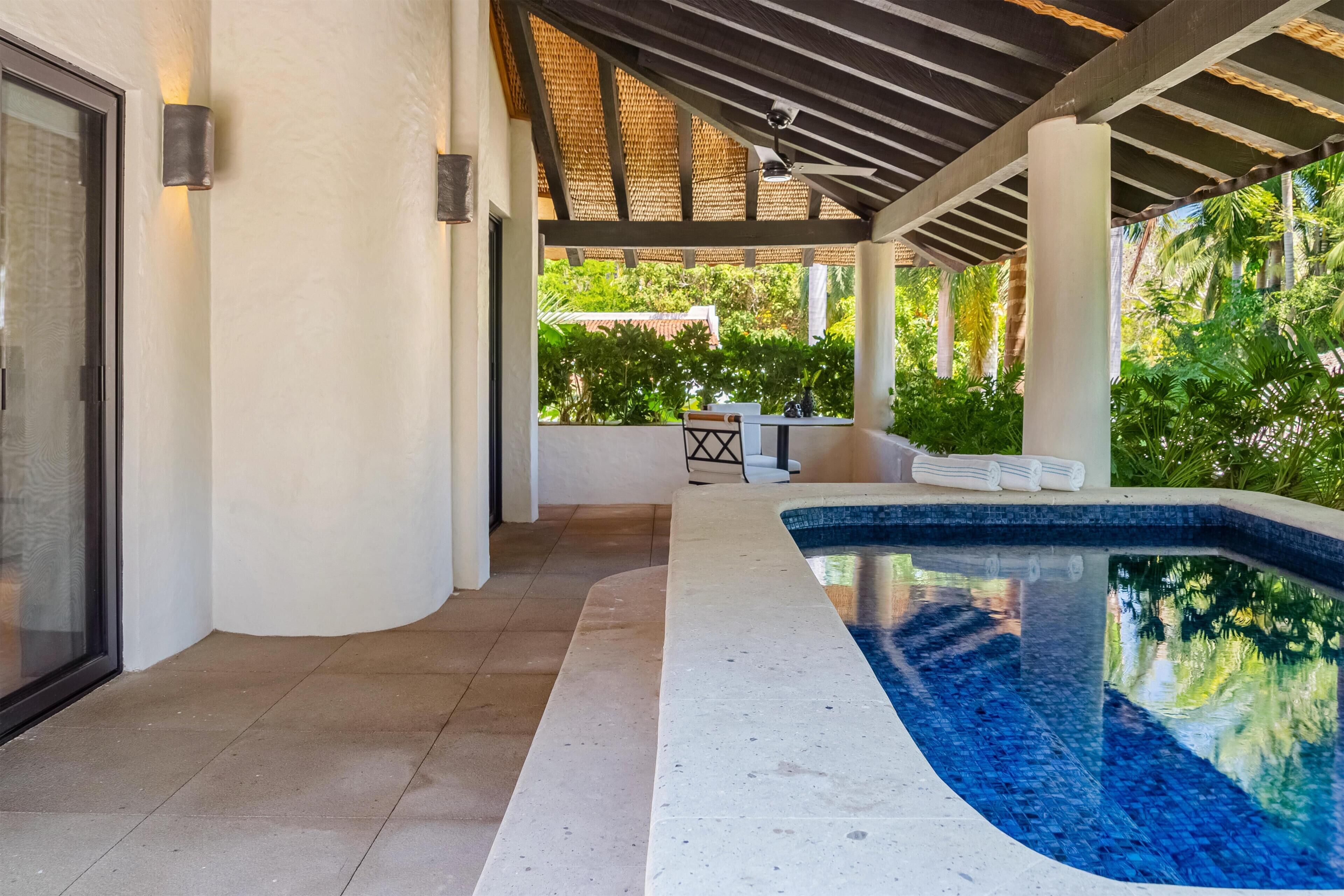 Una piscina exterior techada con una pérgola, un área de descanso y vistas a exuberante vegetación.