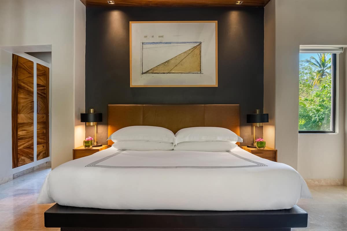 A large bed with a wooden headboard, two bedside tables with lamps, a framed artwork above the bed, and a window with a view of greenery.