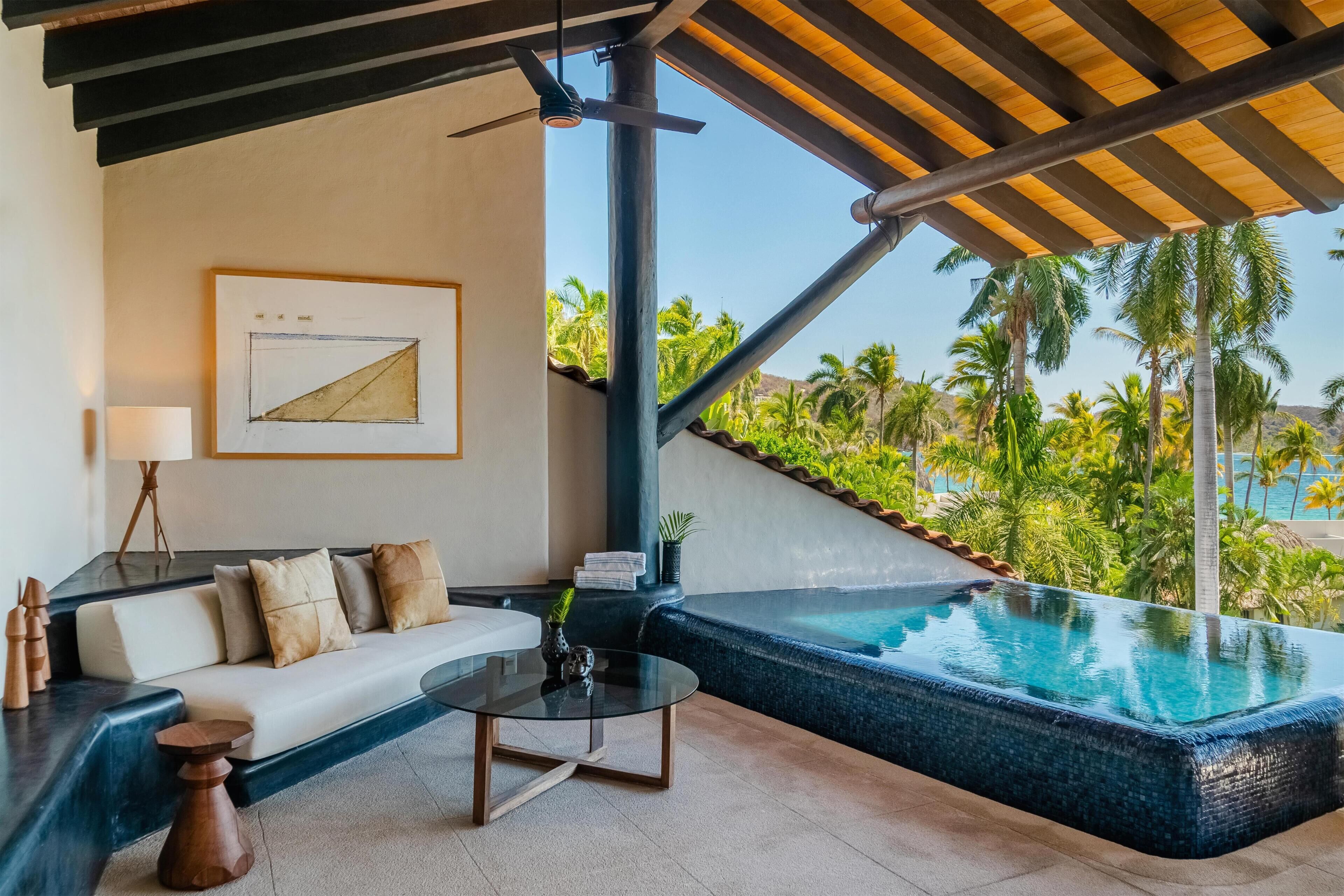 A modern room with a pool, a sofa, a coffee table, and a view of palm trees and a beach.