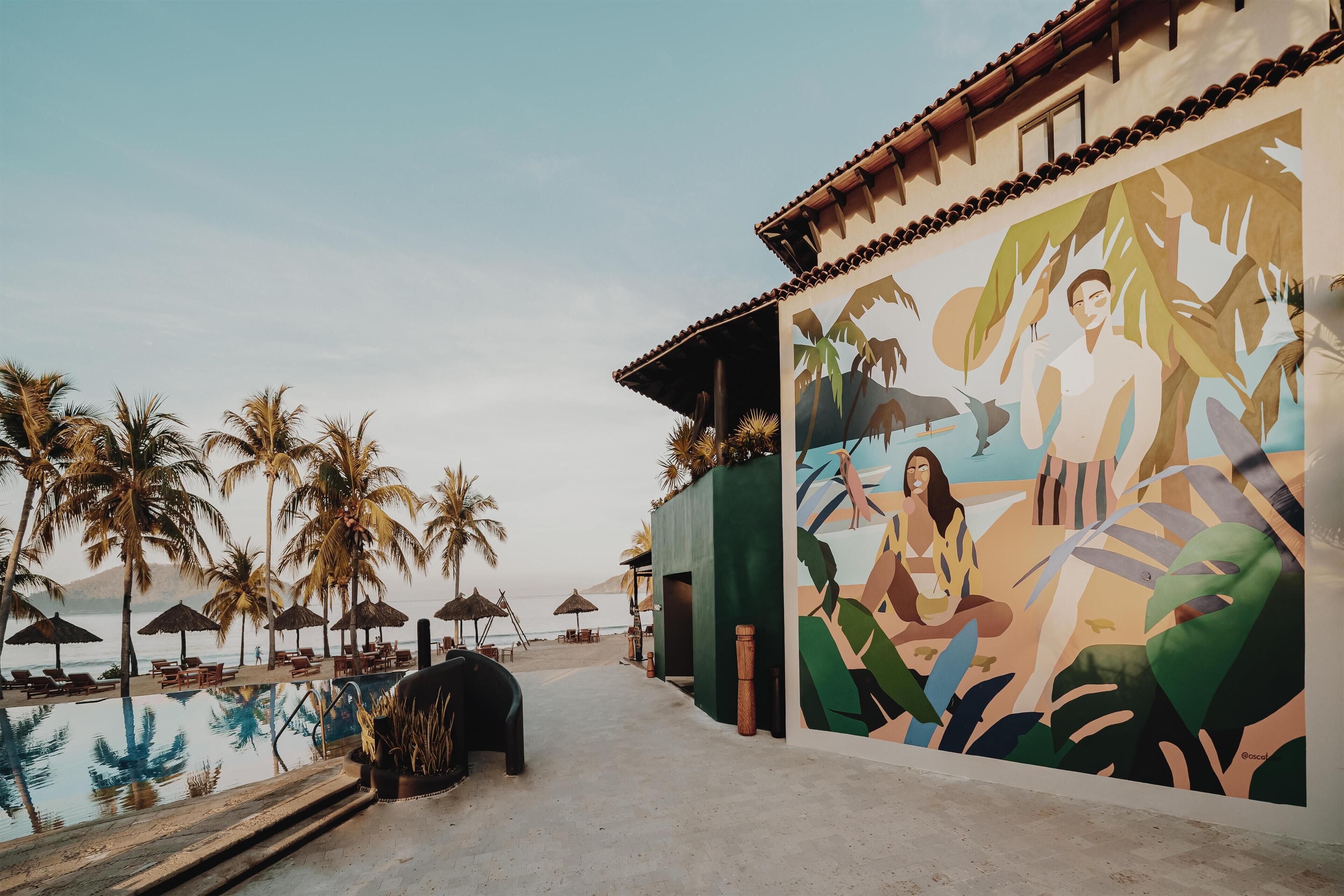 Zona de piscina con un mural, palmeras y vista al océano.