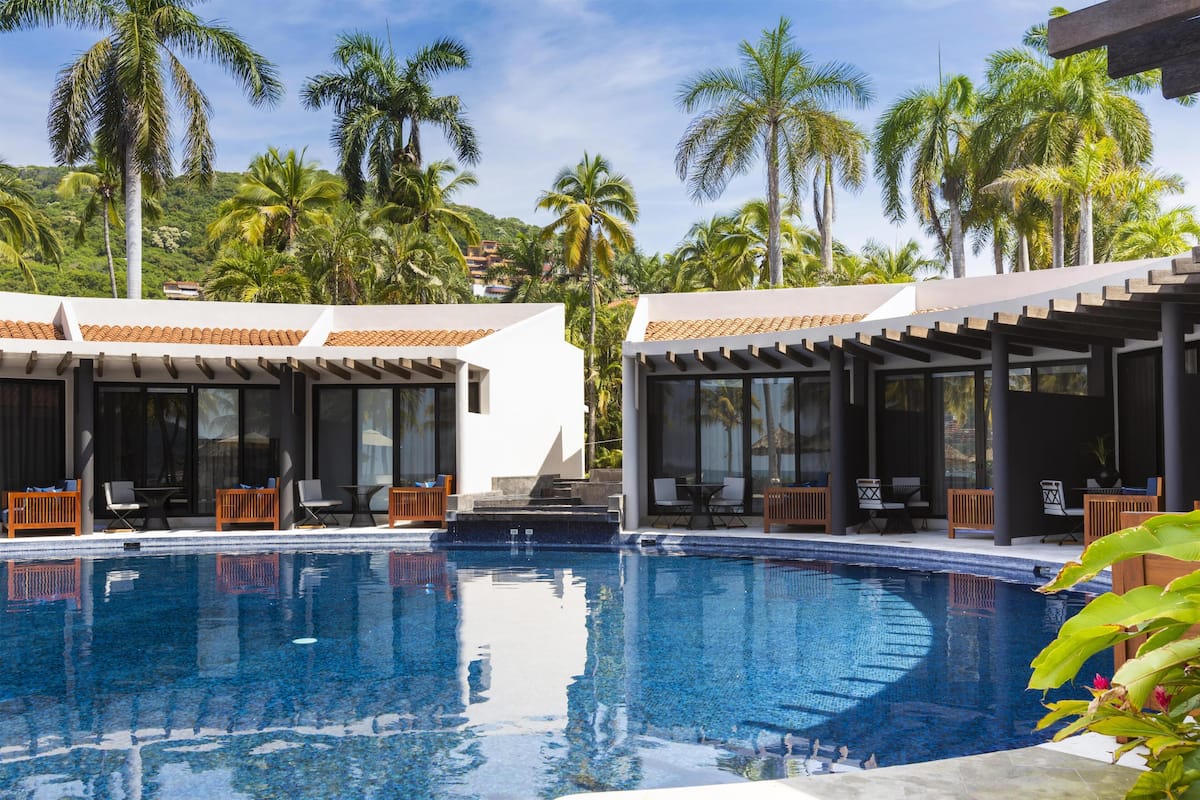 A pool with lounge chairs and umbrellas, surrounded by palm trees and modern architecture.
