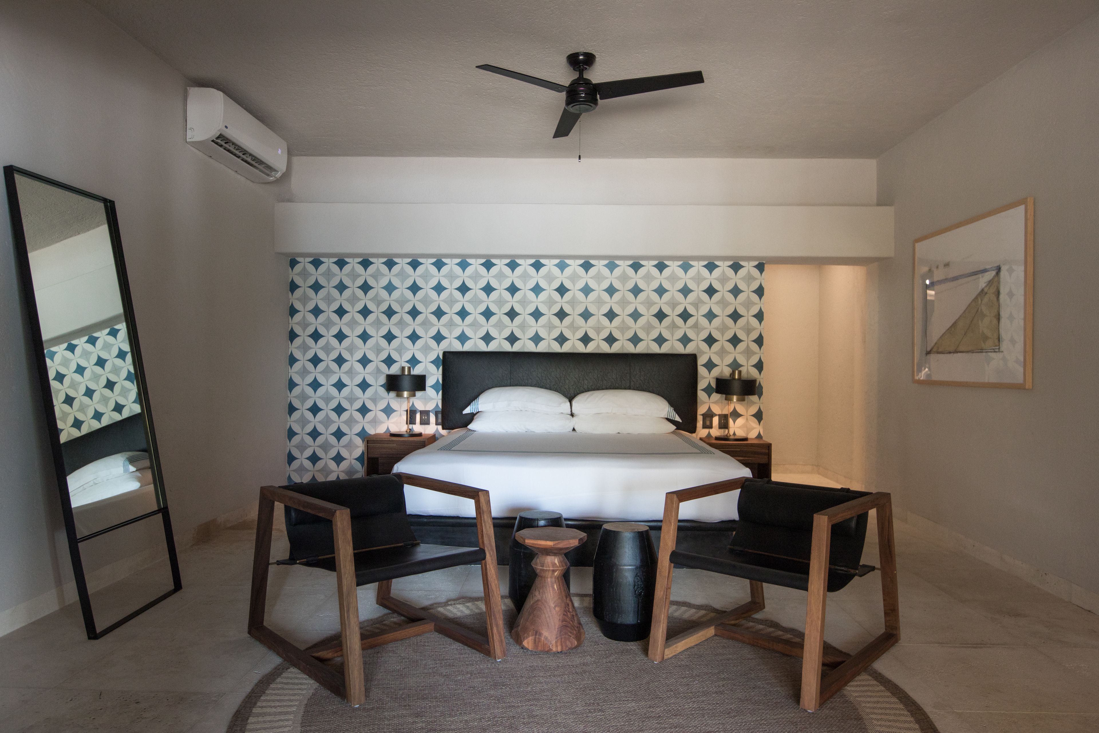 Un dormitorio con cama, ventilador de techo, mesita de noche, espejo y pared con estampado geométrico.