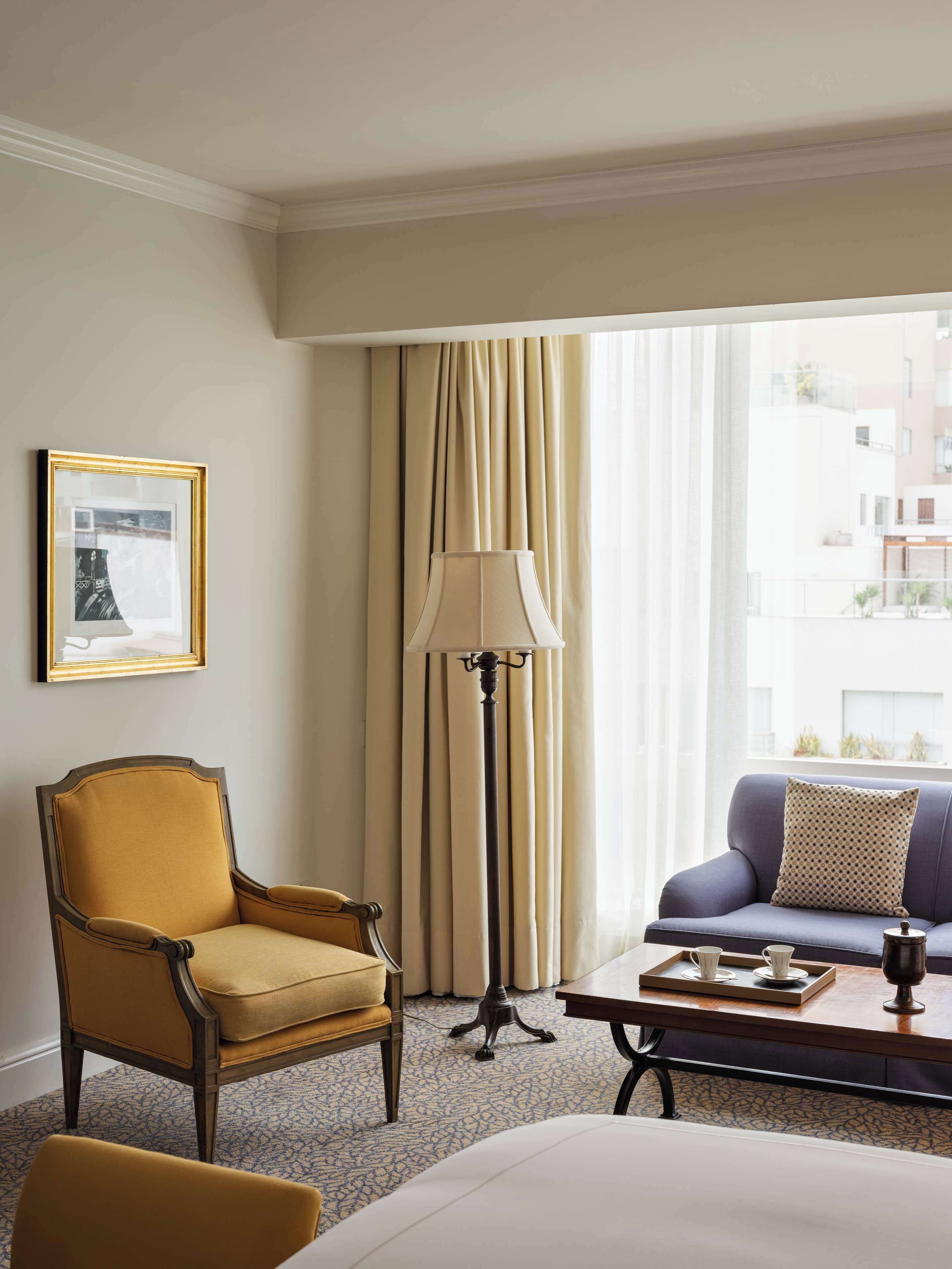 A hotel room with a bed, a sofa, a chair, a floor lamp, a table with a tray, and a framed picture on the wall.