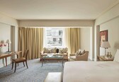A hotel room with a bed, two sofas, a coffee table, a desk, and a chair. There is a window with curtains, a painting on the wall, and a lamp on the desk.