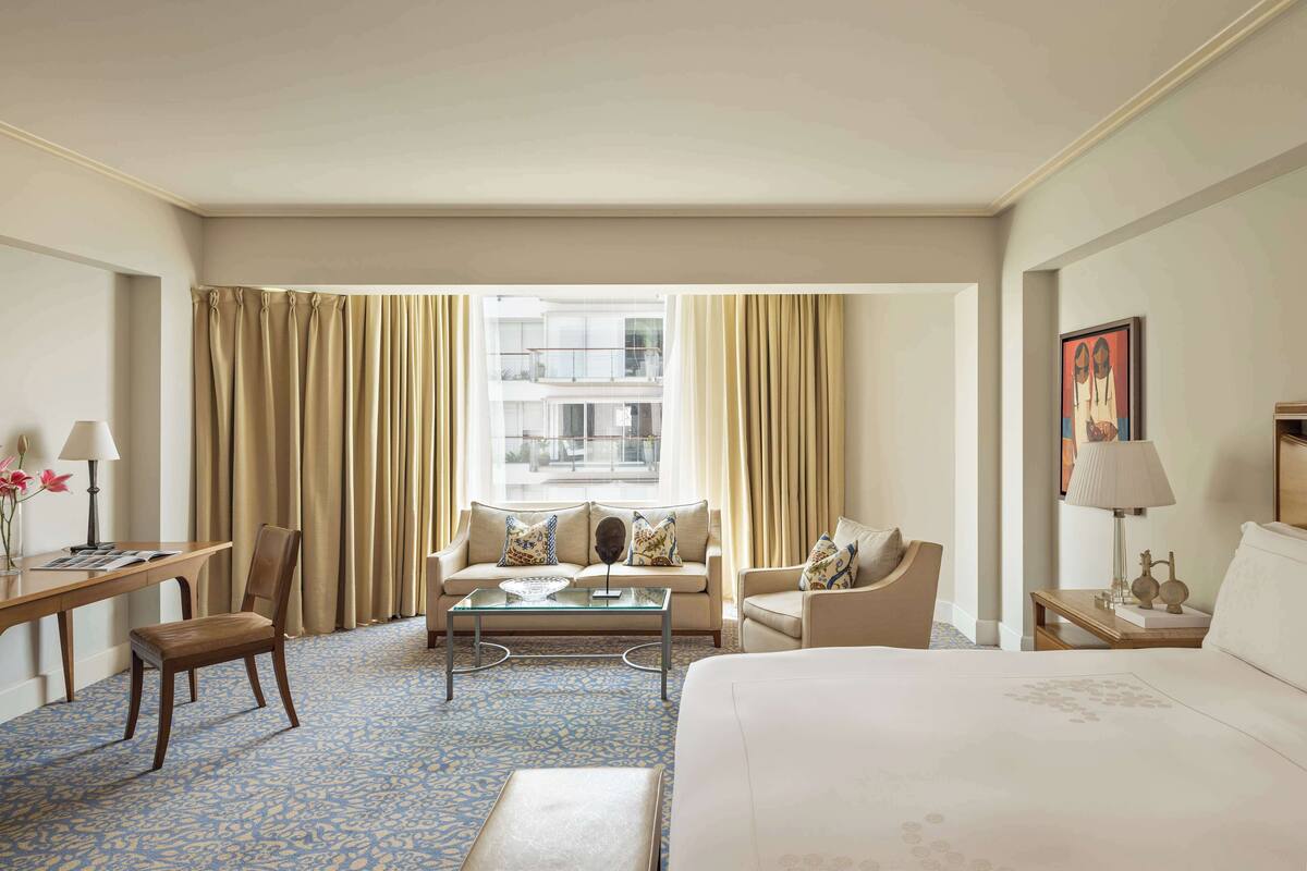 A hotel room with a large bed, a sofa, a coffee table, and a desk. There is a window with curtains, a lamp, and a painting on the wall.
