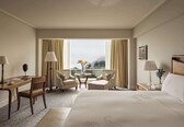 A hotel room with a bed, two armchairs, a desk, a small table with flowers, and a view of the city.