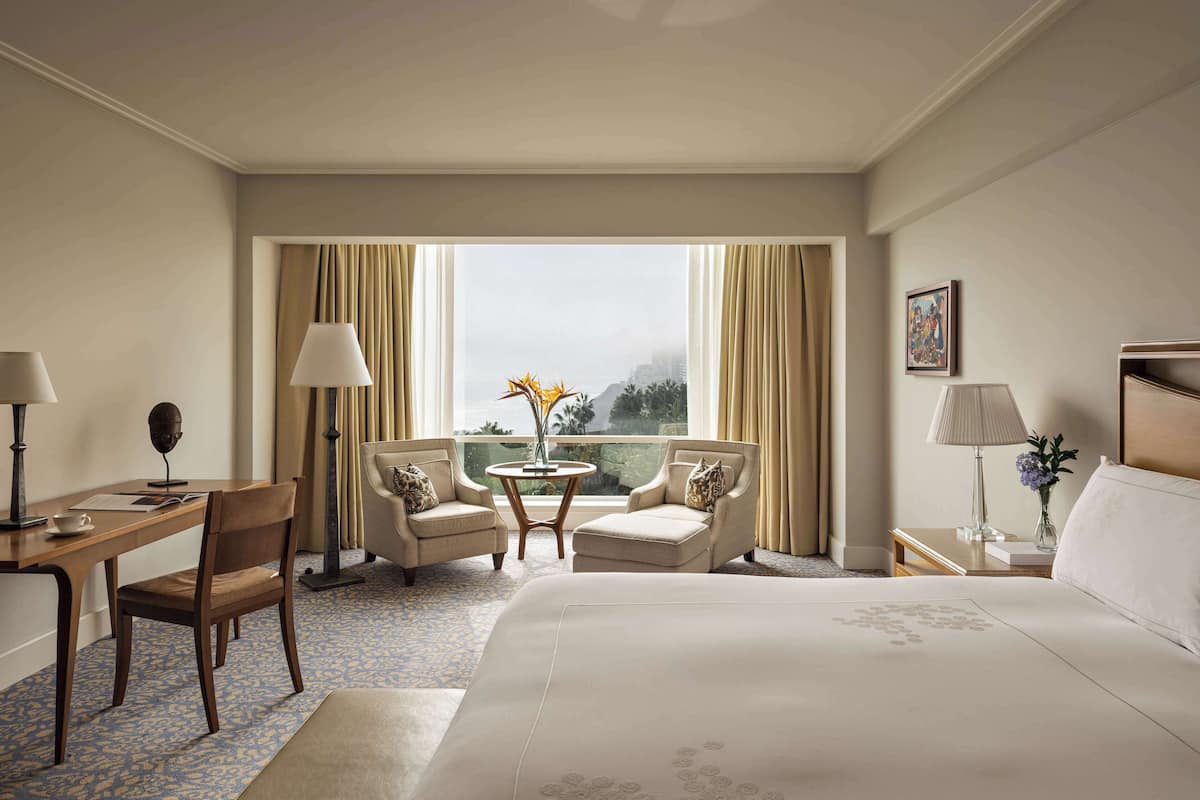 A hotel room with a large bed, two armchairs, a small table with a vase, a desk with a lamp, and a view of the cityscape through large windows.