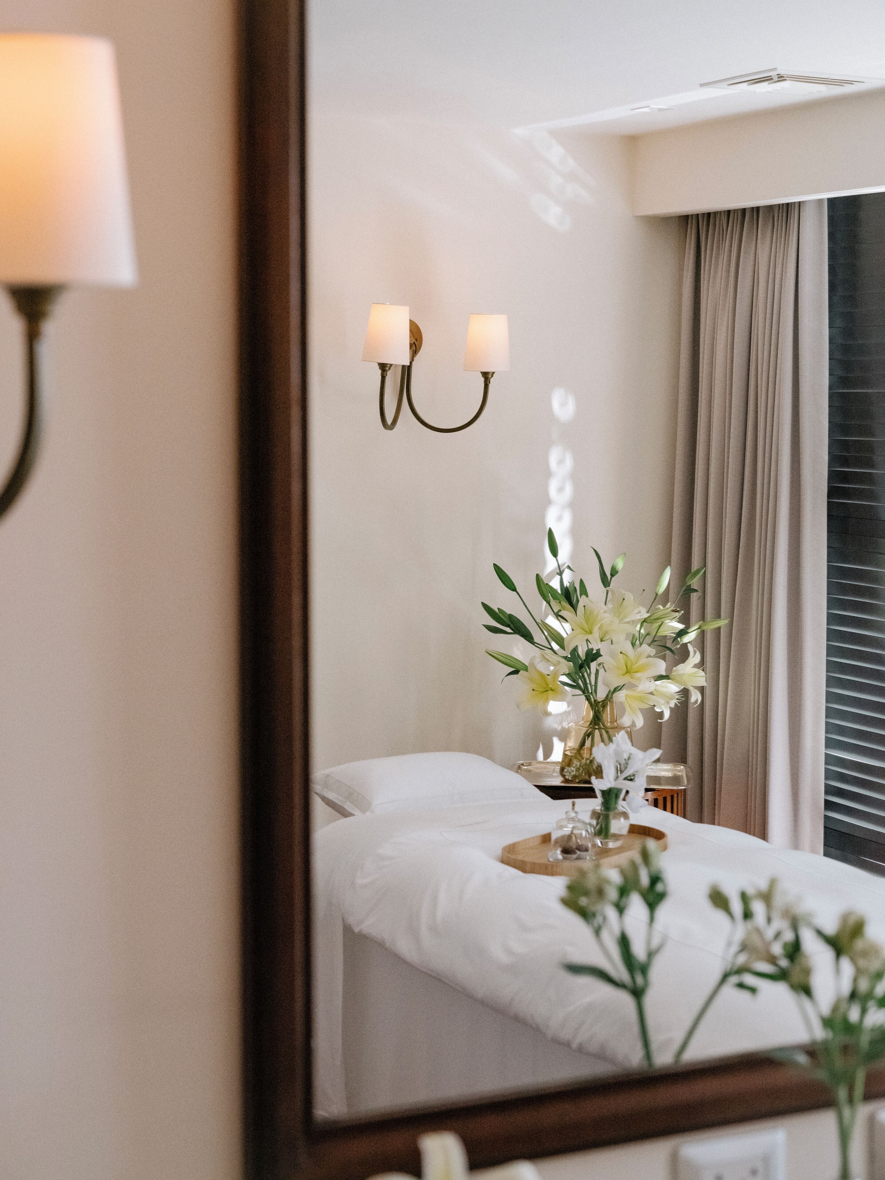 Habitación de hotel con cama, mesita de noche, espejo, lámpara de pared y ventana con cortinas.
