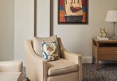 A modern hotel room with a beige armchair, a glass coffee table, a wooden nightstand, and a framed abstract painting on the wall.