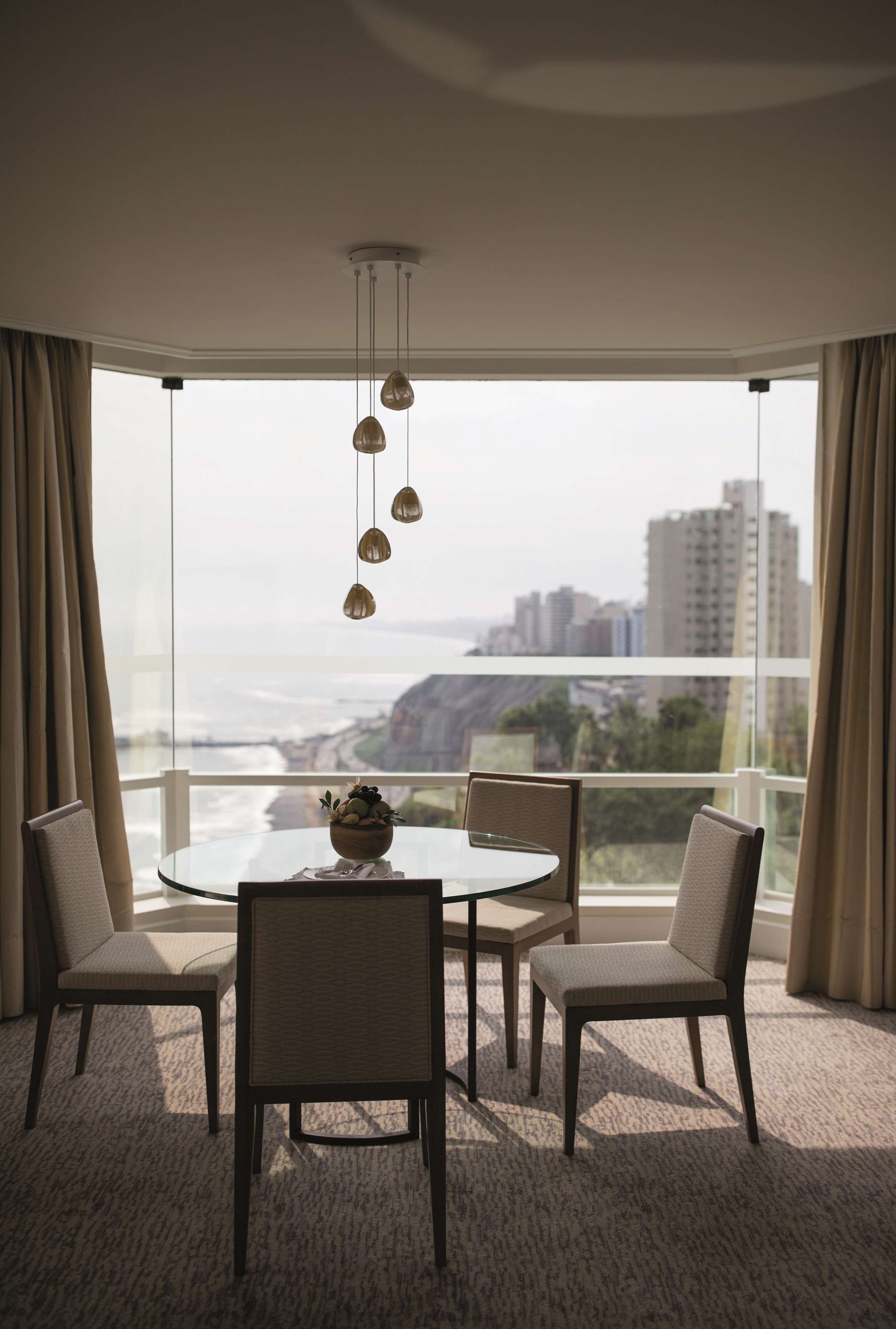 A modern dining room with a glass table and chairs, a hanging light fixture, and a view of the ocean and cityscape.
