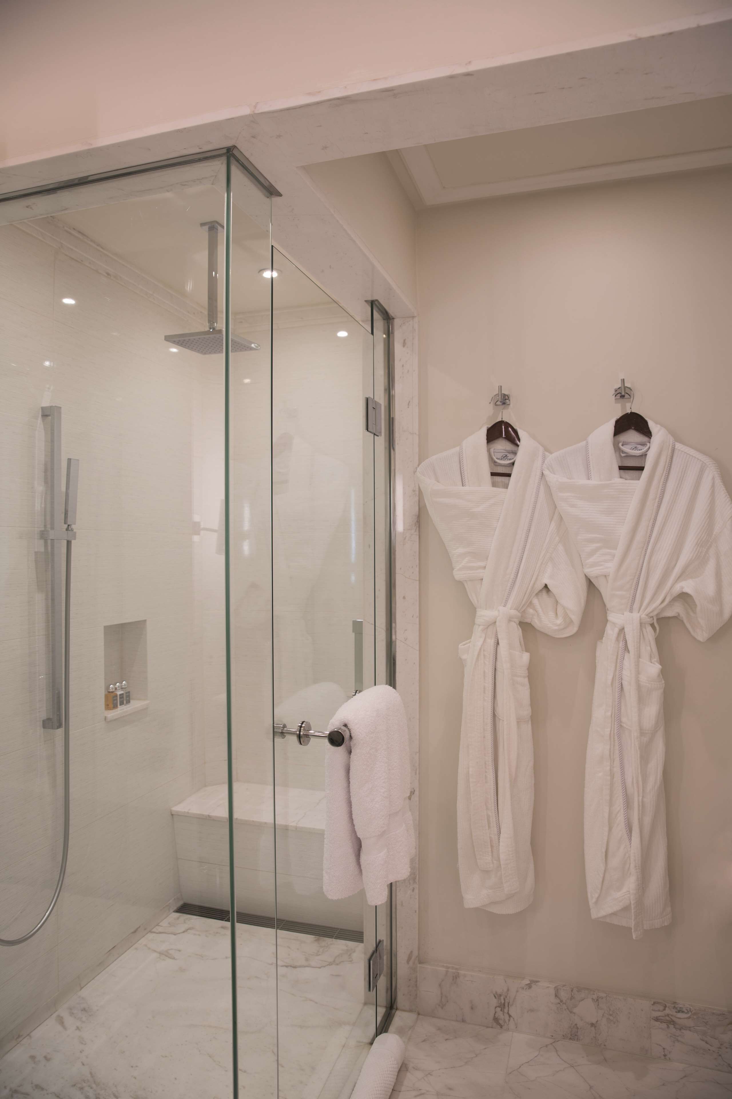 Un baño moderno con ducha de vidrio, suelo de mármol y dos batas blancas colgadas en la pared.