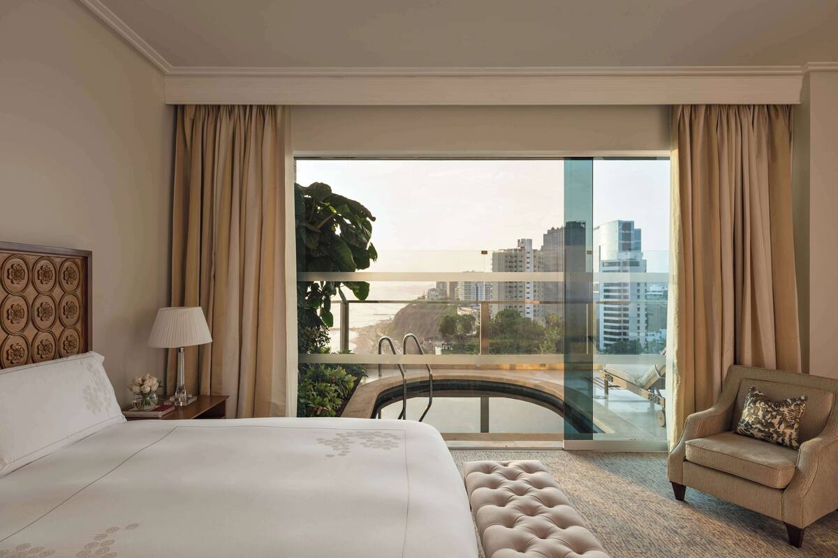A hotel room with a large bed, a sitting area with a chair, a view of a cityscape through a glass wall, and a balcony with a railing.