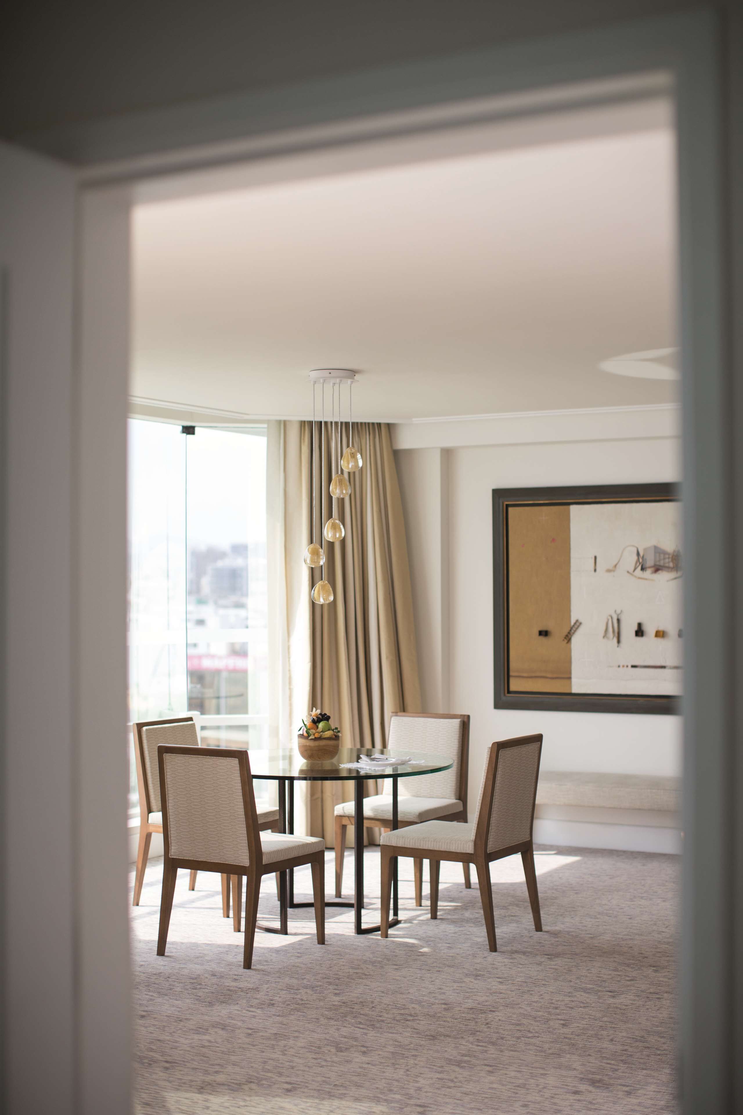 A modern dining area with a glass table, four chairs, a chandelier, and a large window with curtains.