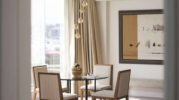 A modern dining area with a glass table, four chairs, a chandelier, and a large window with curtains.