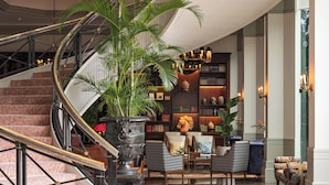 A grand lobby with a curved staircase, a large potted plant, and a bookshelf filled with books and decorative items.