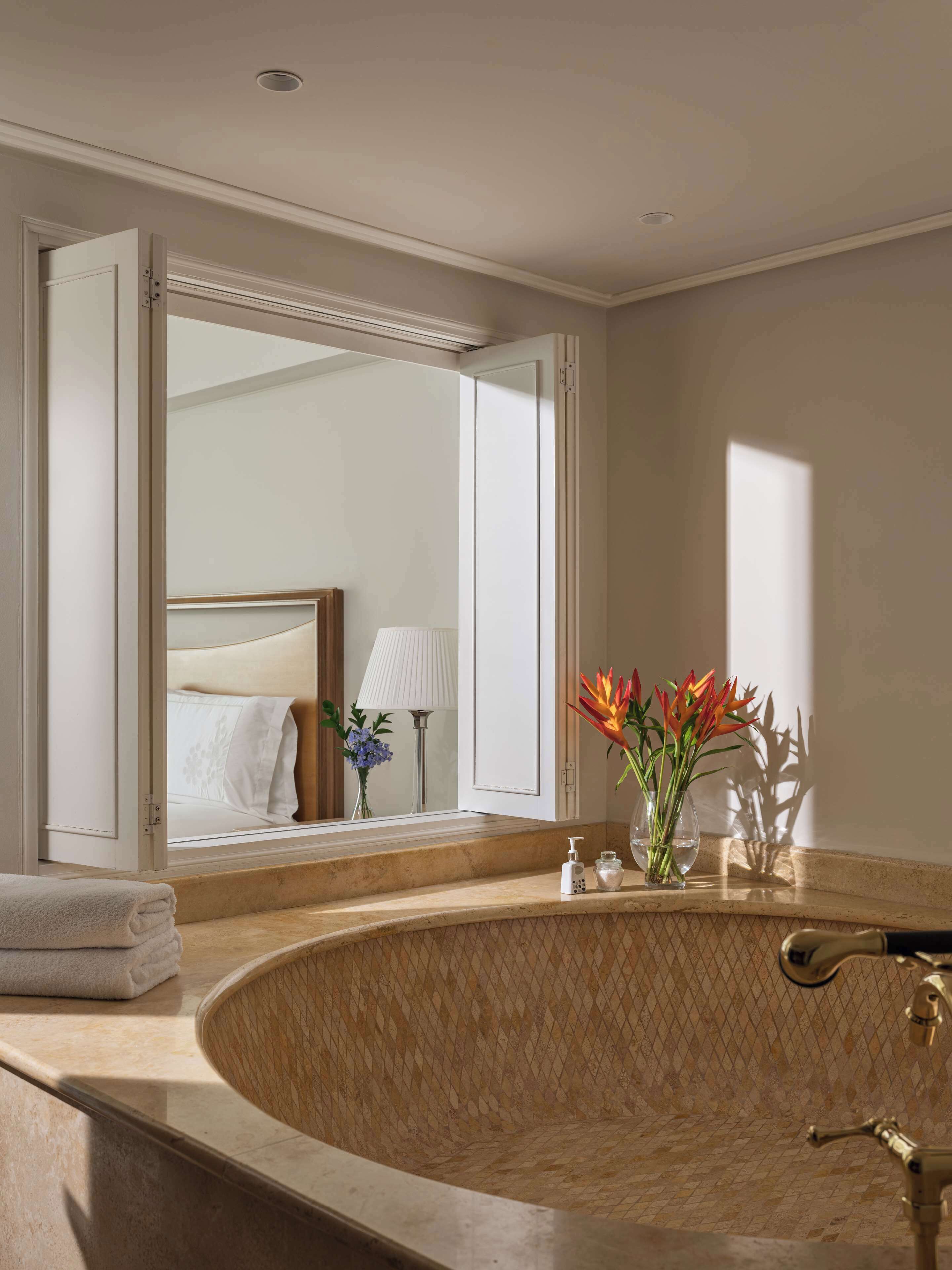 Un baño con una bañera grande, un espejo, una toalla, un florero con flores y una cama visible a través de una puerta abierta.