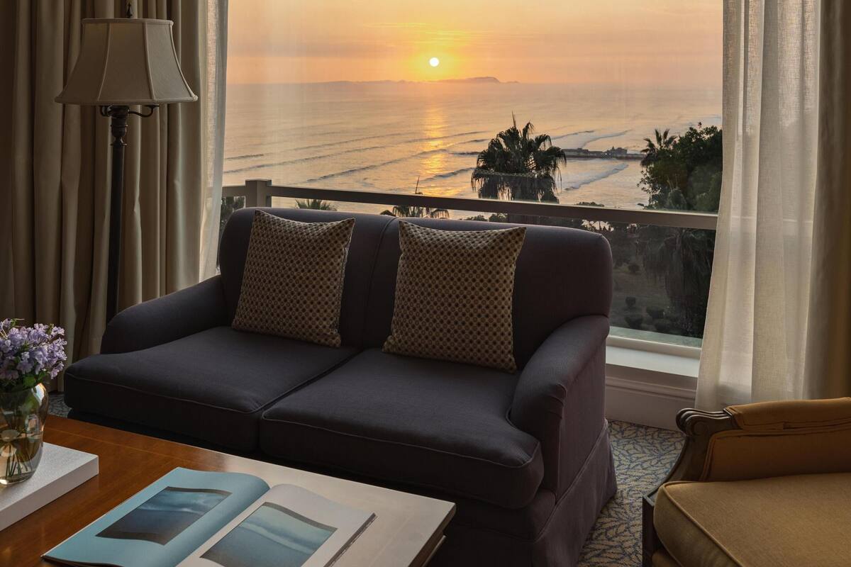 A hotel room with a large window, a sofa, a lamp, and a table with a book.